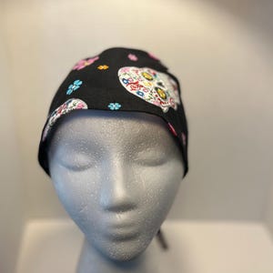 May include: Black skull cap featuring a colorful sugar skull design with floral accents. The cap is adorned with pink, blue, and white flowers. The skull design is centered on the cap, creating a festive and eye-catching look.