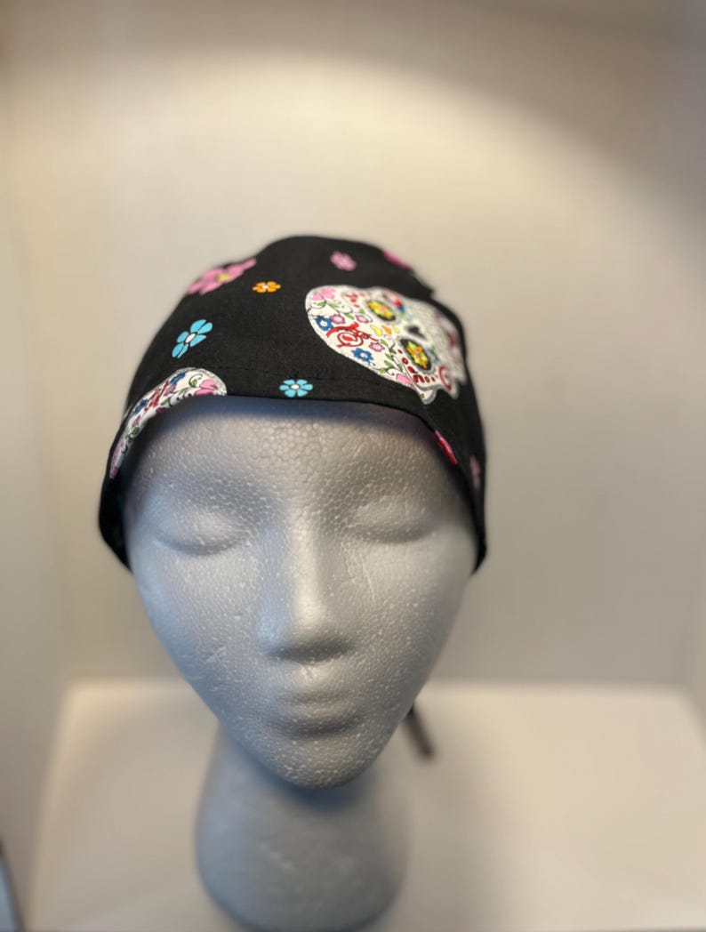 May include: Black skull cap featuring a colorful sugar skull design with floral accents. The cap is adorned with pink, blue, and white flowers. The skull design is centered on the cap, creating a festive and eye-catching look.