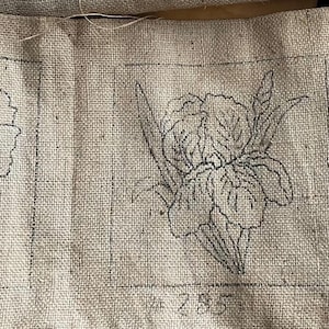 May include: A piece of beige fabric with three floral designs outlined in black ink. The designs include a poppy, an iris, and a rose, each within a rectangular frame. The fabric has a textured appearance.