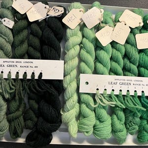 May include: An assortment of green yarn skeins, ranging from light to dark shades, arranged in a tray. The yarn is labeled with tags and color swatches, including "Sea Green" and "Leaf Green" from Appleton Bros. London. Ideal for knitting and crochet projects.