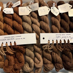 May include: A tray of brown yarn skeins with attached tags labeled with numbers. The yarn colors range from light tan to dark brown. Two cards read "APPLETON BROS. LONDON" with "GOLDEN BROWN" and "FAWN" printed on them.