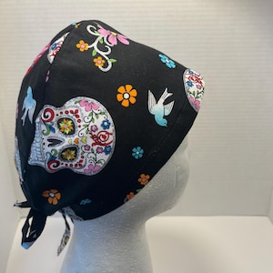 May include: Black surgical scrub cap with a colorful sugar skull and floral pattern. The cap features a tie-back design and is adorned with flowers, birds, and decorative elements in shades of pink, orange, blue, and white.