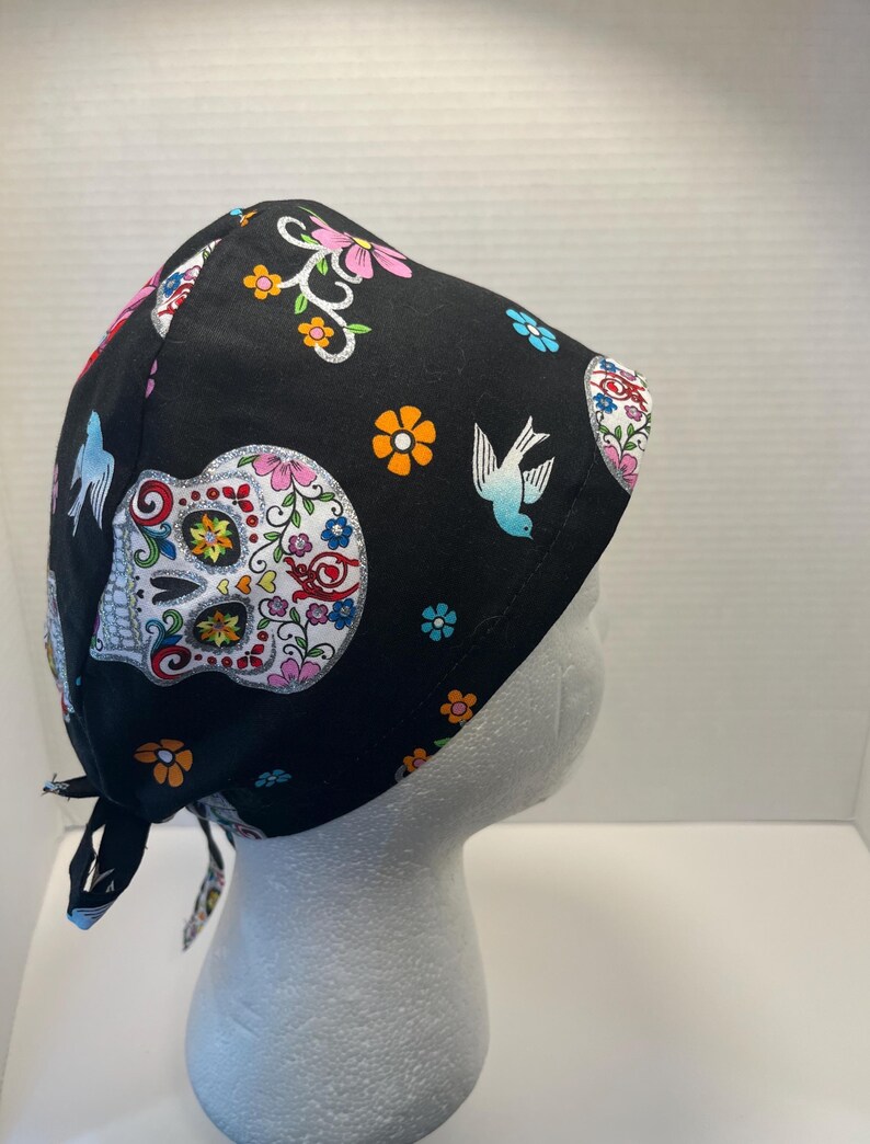 May include: Black surgical scrub cap with a colorful sugar skull and floral pattern. The cap features a tie-back design and is adorned with flowers, birds, and decorative elements in shades of pink, orange, blue, and white.