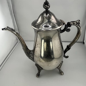 Può includere: Una caffettiera in metallo argentato con un corpo alto e lucido, un beccuccio curvo con dettagli ornamentali e un coperchio abbinato con un pomello decorativo. Il recipiente poggia su tre piccoli piedini scolpiti e ha un manico curvo.