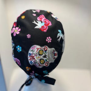 May include: Black surgical scrub cap with a colorful Day of the Dead sugar skull, flowers, and birds pattern. The cap has a tie closure and is made of a soft, patterned fabric. The design includes pink, blue, and yellow floral elements.