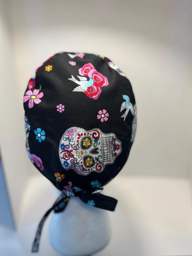 May include: Black surgical scrub cap with a colorful Day of the Dead sugar skull, flowers, and birds pattern. The cap has a tie closure and is made of a soft, patterned fabric. The design includes pink, blue, and yellow floral elements.