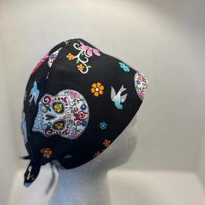 May include: Black skull cap with a colorful sugar skull design, flowers, and birds. The cap is made of fabric and has ties in the back. The sugar skull has a white base with colorful floral accents.