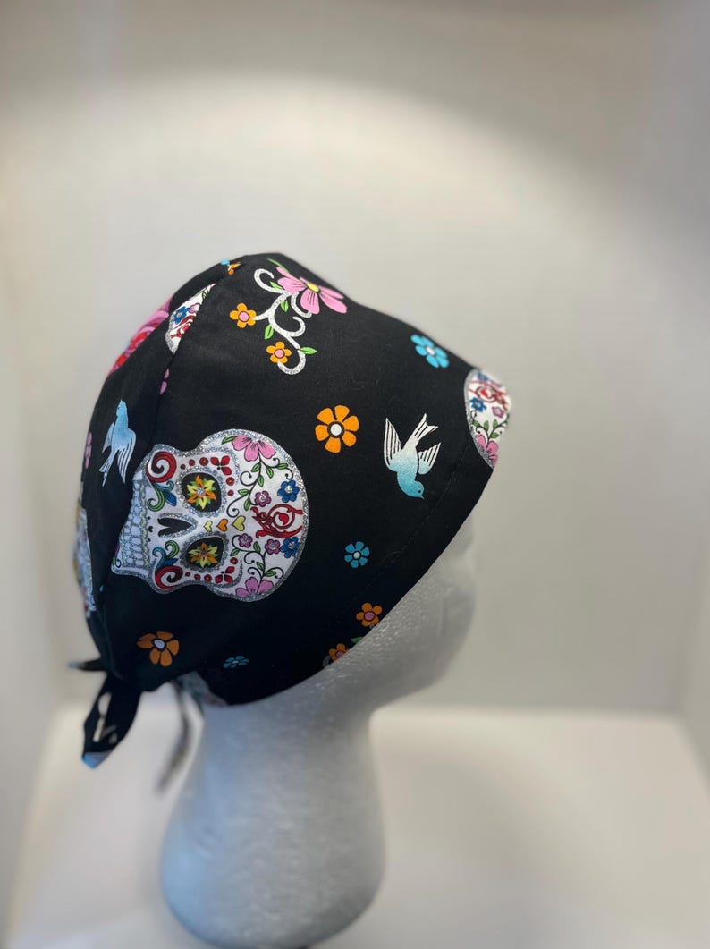 May include: Black skull cap with a colorful sugar skull design, flowers, and birds. The cap is made of fabric and has ties in the back. The sugar skull has a white base with colorful floral accents.