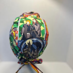May include: A patterned scrub cap with a stained-glass effect, depicting characters and scenes from a fantasy series. The cap, with a tie closure, is made of fabric in shades of green, yellow, red, and blue.