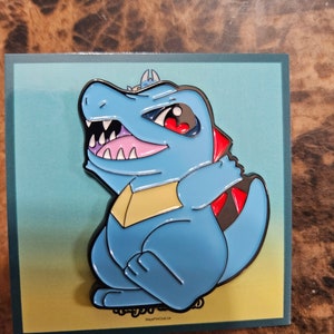 May include: A blue enamel pin depicting a cartoon alligator-like creature with a fierce expression. The pin has a black outline, red accents, and a yellow chest. The creature is set against a gradient background.