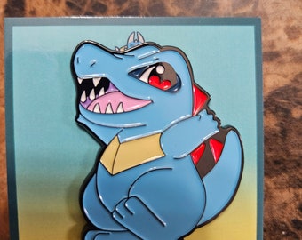 Puede incluir: Un pin de esmalte azul con una criatura de dibujos animados parecida a un caimán con una expresión feroz. El pin tiene un contorno negro, detalles en rojo y un pecho amarillo. La criatura está sobre un fondo degradado.