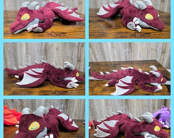 May include: Plush dragon toy in various poses. The dragon is primarily burgundy with gray accents on the wings, horns, and claws. The dragon has a yellow eye with a green outline. The plush toy is lying down on a wooden surface.