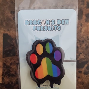 May include: A black phone grip shaped like a paw print, featuring a rainbow design. The grip is on a light blue card with the text "DRAGON'S DEN FURSUITS". The packaging is clear plastic.