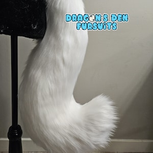 May include: White fluffy faux fur tail for cosplay or costume. The tail is curled at the end and is attached to a black stand. The text "DRAGONS DEN FURSUITS" is visible in the image.