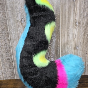 May include: A vibrant, curved faux fur tail. The tail is primarily black, with neon green and yellow patches. It transitions to a bright blue, with a pink stripe and a blue tip. The tail is designed to be worn as part of a costume.