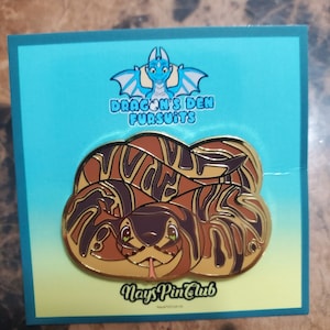 May include: A gold-coloured enamel pin featuring a cartoon snake with brown, orange and yellow stripes. The snake has green eyes and a pink tongue. The pin is on a blue background with the text "Dragon's Den Furs uits" and "Nays Pin Club".