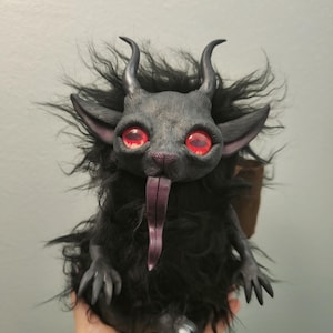 Puede incluir: Una criatura negra y peluda con cuernos, ojos rojos y una larga lengua morada. La criatura está sentada en una mano.