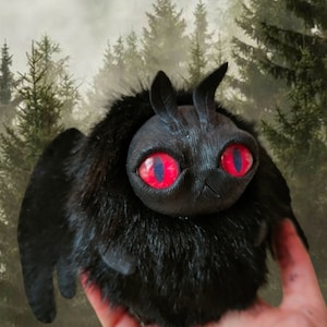 Può includere: Peluche nero con grandi occhi rossi e una texture pelosa. Il giocattolo ha un paio di piccole corna e ali. Il giocattolo è tenuto in una mano contro uno sfondo sfocato di una foresta.