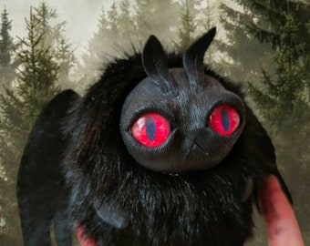 May include: Black plush toy with large red eyes and a furry texture. The toy has a pair of small horns and wings. The toy is being held in a hand against a blurry background of a forest.