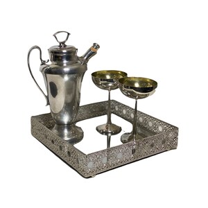 May include: A silver cocktail shaker, two champagne coupe glasses, and a decorative tray. The tray has a mirrored surface and an openwork design. The glasses have gold-coloured interiors.