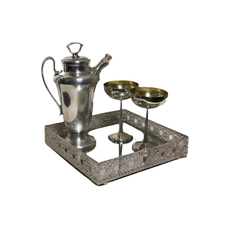 May include: A silver cocktail shaker, two champagne coupe glasses, and a decorative tray. The tray has a mirrored surface and an openwork design. The glasses have gold-coloured interiors.