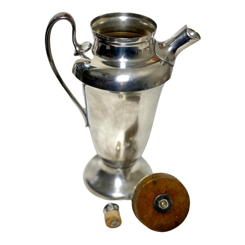 May include: A vintage silver cocktail shaker with a handle and spout. The shaker has a bulbous base and a lid with a wooden top. A small cork stopper is also present. The items are displayed on a white background.