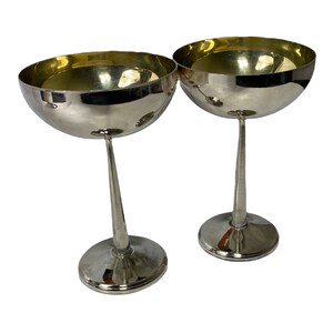 May include: Two silver champagne coupe glasses. Each glass has a rounded bowl with a gold-coloured interior and a slender stem. The glasses are displayed on a white background.