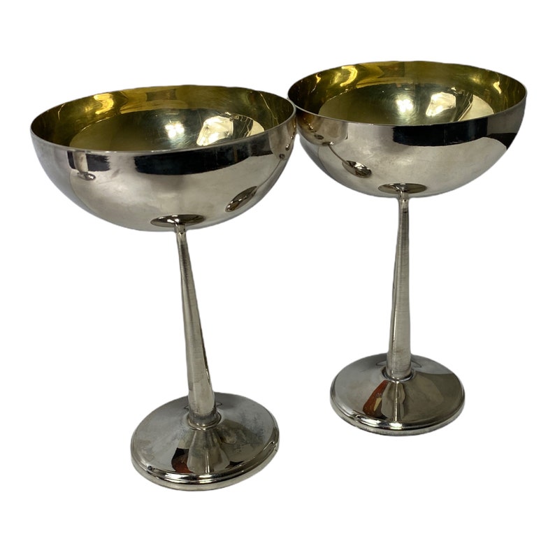 May include: Two silver champagne coupe glasses. Each glass has a rounded bowl with a gold-coloured interior and a slender stem. The glasses are displayed on a white background.