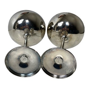 May include: Two silver-coloured wine glasses with a spherical bowl, stem, and circular base. The glasses are reflective, with the base and stem appearing to be made of the same material. The glasses are set against a white background.