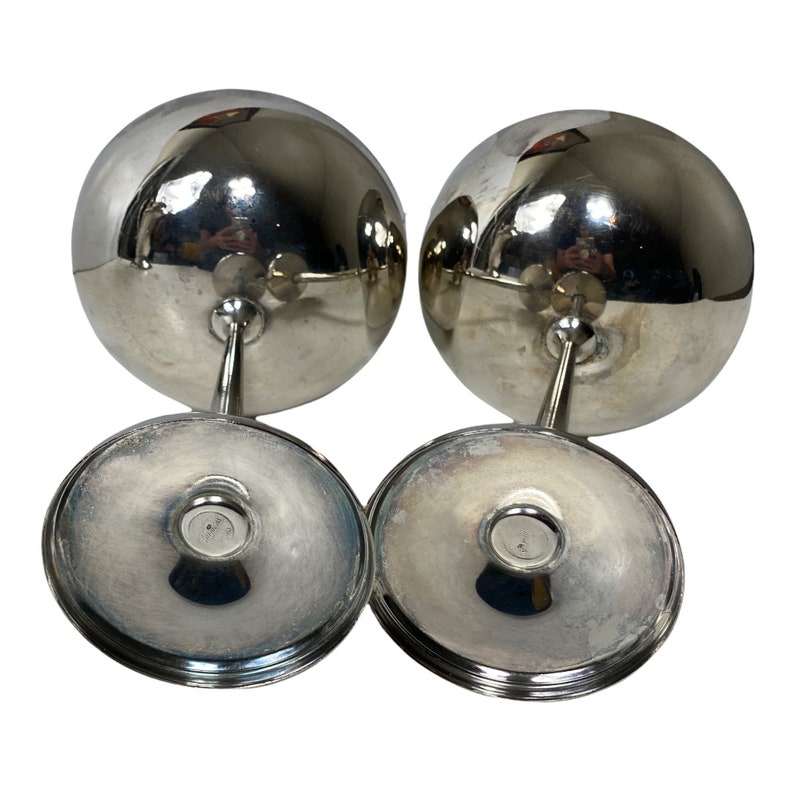 May include: Two silver-coloured wine glasses with a spherical bowl, stem, and circular base. The glasses are reflective, with the base and stem appearing to be made of the same material. The glasses are set against a white background.