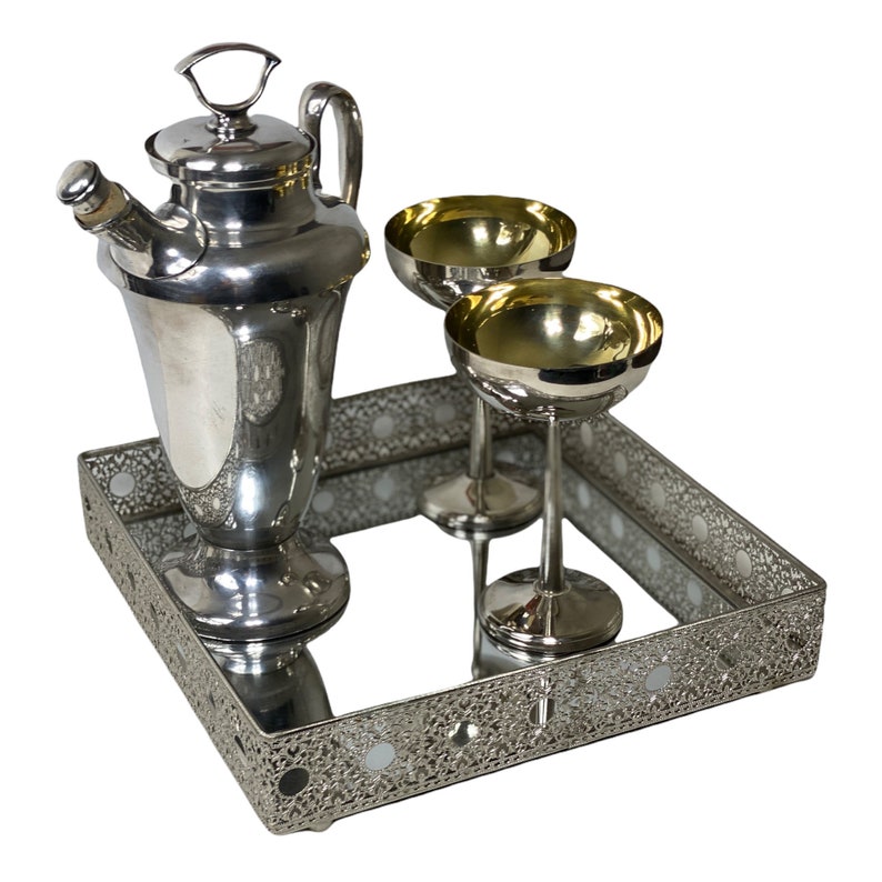 May include: A silver cocktail set on a mirrored tray. The set includes a cocktail shaker, two coupe glasses, and a decorative tray with an openwork design. The glasses have gold-coloured interiors.