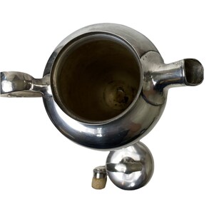 May include: A silver teapot with a rounded body, spout, and handle. The lid and a small stopper are next to the teapot. The interior of the teapot is visible, showing a dark interior. The teapot is on a white background.
