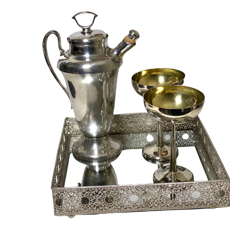 May include: A silver cocktail shaker with a handle, a cork stopper, and two champagne coupe glasses sit on a mirrored tray with a decorative, openwork border. The glasses have gold-coloured interiors.