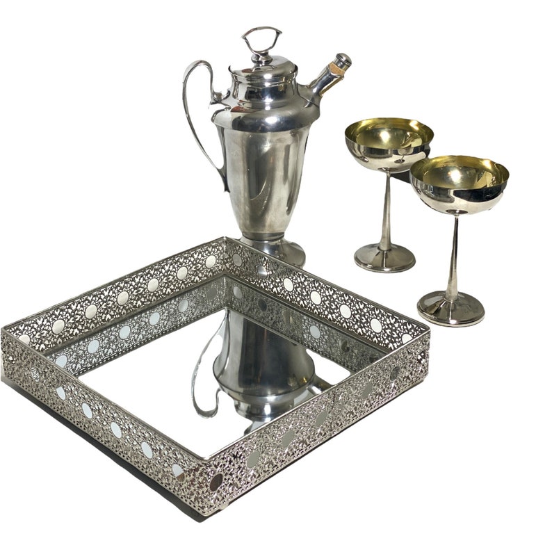 May include: A silver cocktail set comprising a cocktail shaker, two champagne coupe glasses, and a decorative tray. The tray has a mirrored base and an ornate, openwork border. The glasses and shaker have a polished finish.