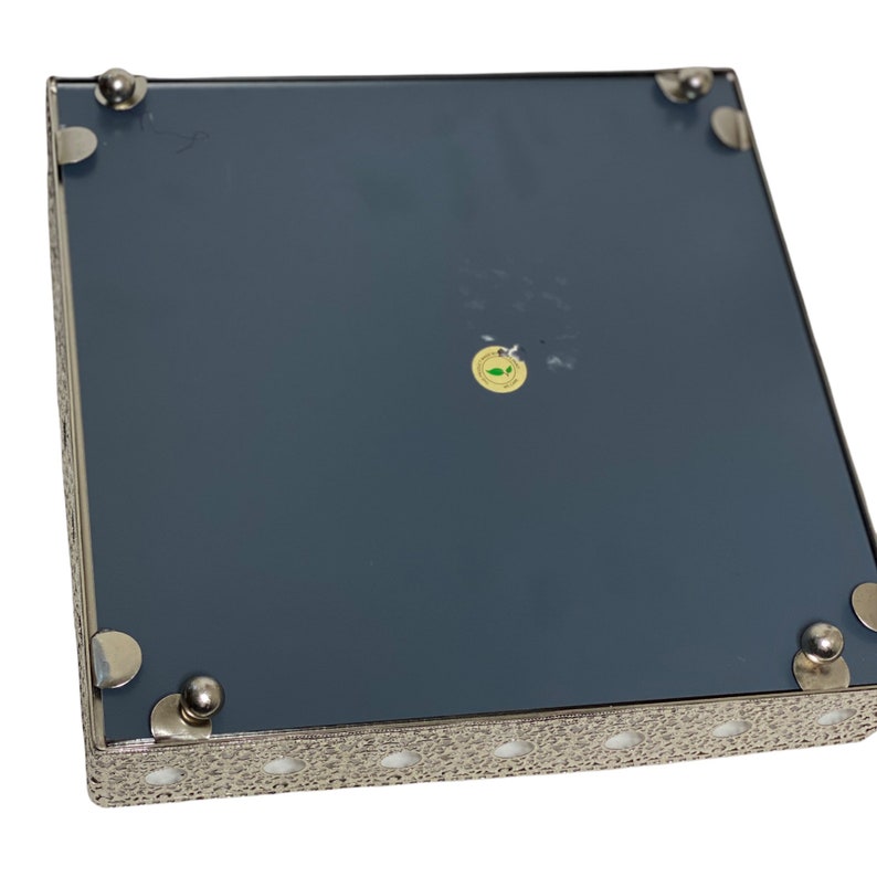 May include: A square decorative tray with a dark blue surface and a silver filigree border. The tray has small, silver ball feet and corner accents. A small, round sticker is visible on the surface.