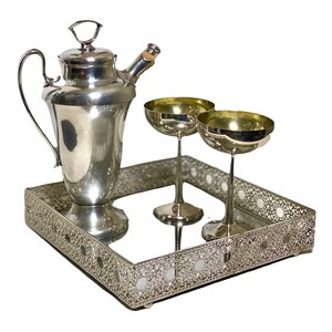 May include: A silver cocktail set comprising a shaker, two coupe glasses, and a decorative tray. The tray features a mirrored surface and an ornate, openwork design. The glasses have gold-coloured interiors.