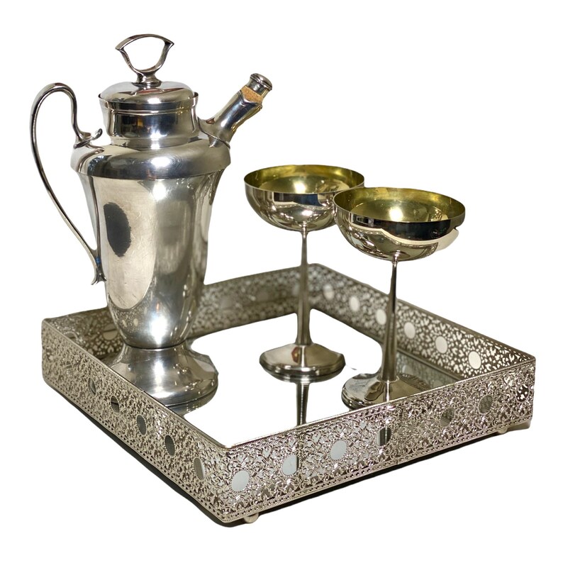 May include: A silver cocktail set comprising a shaker, two coupe glasses, and a decorative tray. The tray features a mirrored surface and an ornate, openwork design. The glasses have gold-coloured interiors.