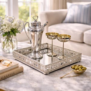 May include: A silver cocktail shaker, two gold-rimmed champagne glasses, and a decorative silver tray with a mirrored surface. The tray holds the glasses and shaker, with a small bowl of olives nearby. A vase of flowers sits in the background.