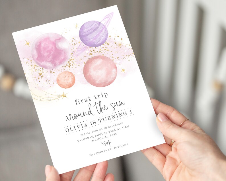 May include: A pink and purple watercolour illustration of planets and stars with gold glitter. The text reads "first trip around the sun Olivia is turning 1" with details for a birthday party.