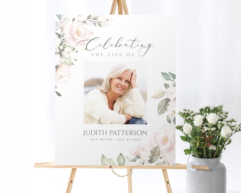 May include: A white memorial sign with a watercolor floral design featuring pink roses and green leaves. The sign includes a photo of a woman, the text "Celebrating the Life of", a name, and dates.