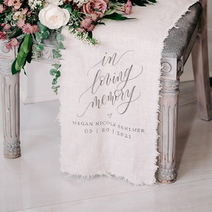 May include: A white linen chair sash with the text "in loving memory of MEGAN NICOLE BEHLMER 09 | 20 | 2021" printed in black. A bouquet of pink and white flowers sits on the chair.