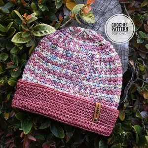 May include: A pink and white crocheted beanie with a brown leather tag that says "S&D". The beanie is sitting on a bed of green leaves.