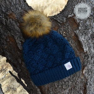 May include: A blue crocheted beanie with a brown faux fur pom pom. The beanie has a textured stitch pattern and a small white label with a pink flower design.