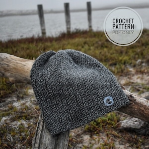 Puede incluir: Un gorro de punto de crochet gris con un patrón de puntes texturizado. El gorro está sentado en una rama con un fondo borroso de un cuerpo de agua y una valla de madera.