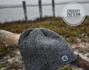 May include: A gray crochet beanie with a textured stitch pattern. The beanie is sitting on a branch with a blurry background of a body of water and a wooden fence.