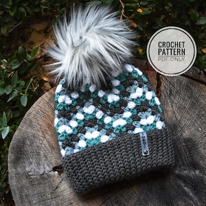 Könnte beinhalten: Gestrickte Mütze mit weißem Bommel, die ein geometrisches Muster in Grün-, Grau- und Weißtönen aufweist. Die Mütze hat einen dunkelgrauen Saum und ein schwarzes Etikett mit dem Text "S&D".