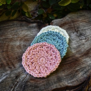 Op de afbeelding: Drie gehaakte make-up remover pads in roze, blauw en wit. De pads liggen op elkaar gestapeld op een houten oppervlak.