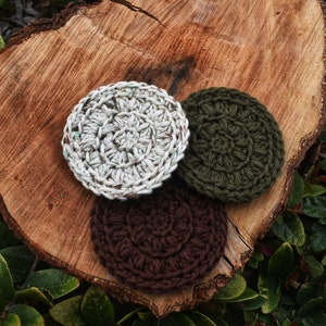 May include: Three crocheted round scrubbers in different colours: white with green flecks, dark green, and brown. The scrubbers are on a wooden surface.