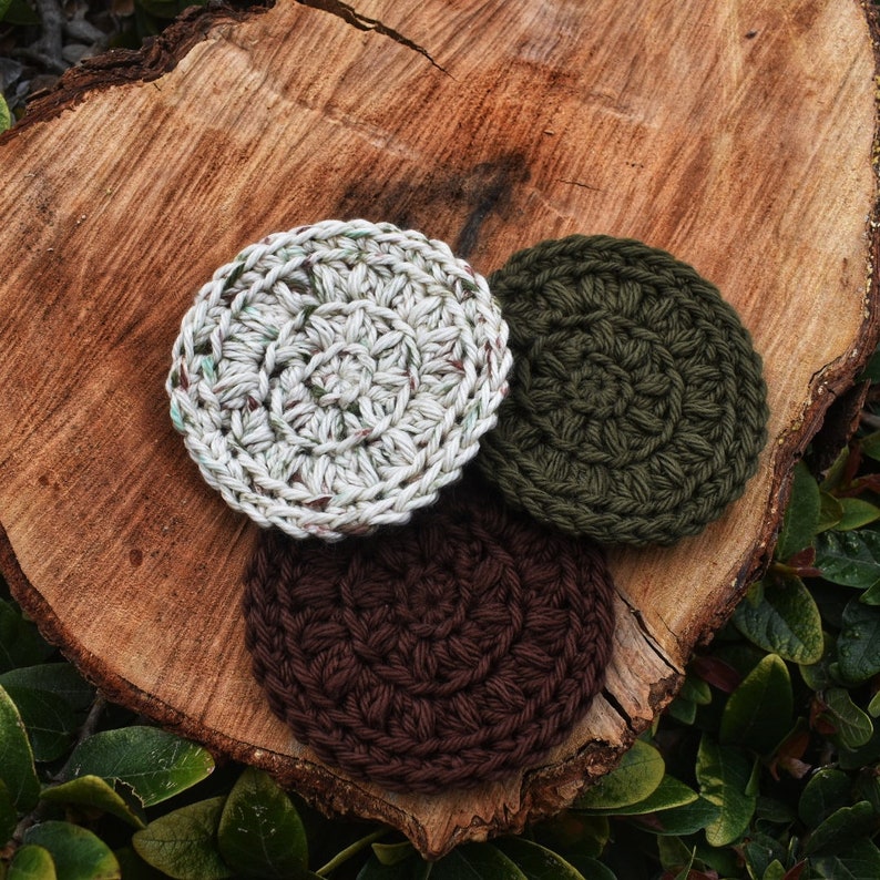 May include: Three crocheted round scrubbers in different colours: white with green flecks, dark green, and brown. The scrubbers are on a wooden surface.