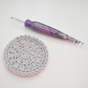 May include: A grey crocheted circle with a purple and silver crochet hook.